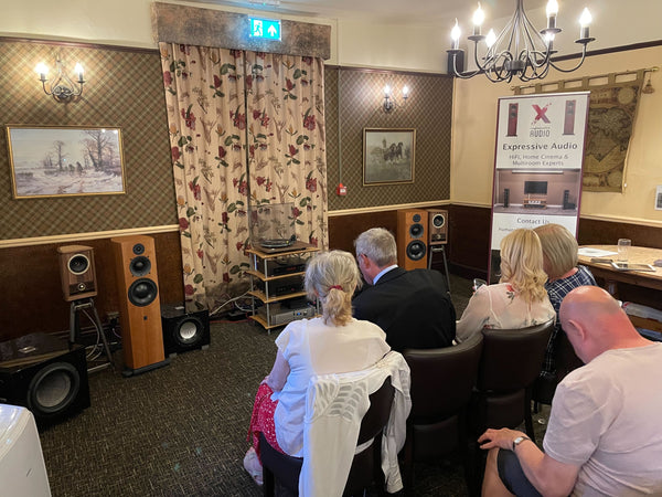 A photo of a HiFi system set up in a room in a pub, and on the right hand side there are some people sitting in chairs listening to the HiFi system.