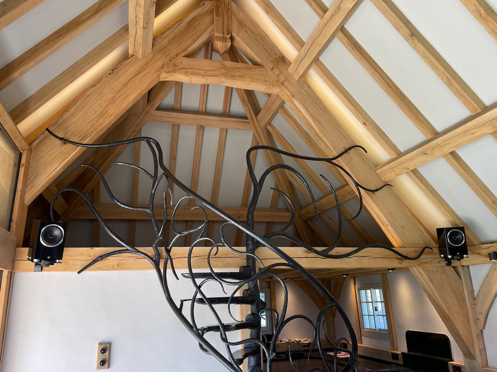 A photo showing a black steel spiral staircase shaped to look like a tree with branches extending out. The staircase goes up to a small loft room area, with exposed light wood beams and eaves against white painted ceiling and walls. Two black loudspeakers have been installed with wall mounts on the beam at the bottom of the loft room.