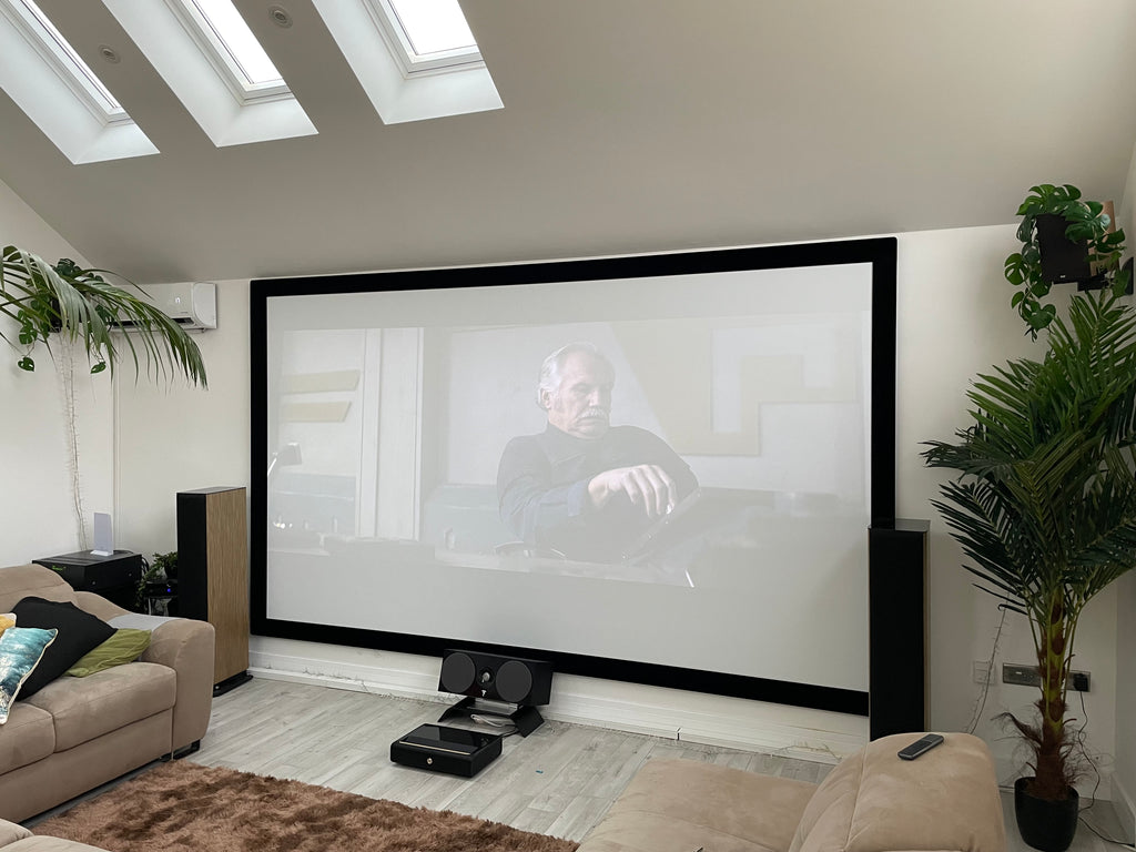 A photo of a big projector screen showing a scene from a film with a man sitting at a desk. The projector screen is on a white wall with a sloped ceiling above it and centre speaker on the floor in front of it, with loudspeakers either side. In the corners of the room on each side of the projector screen are plants. At the bottom of a photo the edges of a sofa are visible in front of the projector screen.