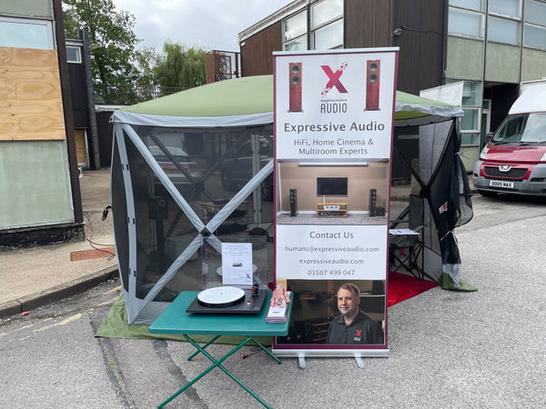 A photo of a green gazebo tent in a car park with a HiFi system set up inside and a banner outside advertising Expressive Audio next to a green folding table with a turntable on top of it.