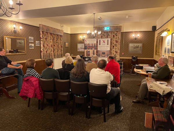 A group of people sitting in rows of chairs in a room at a pub listening to music on a HiFi system which is set up at the front of the room. There are also banners advertising Expressive Audio, a HiFi, Home Cinema & Multiroom retailer.