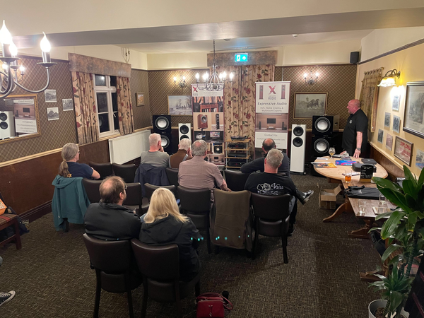 A photo of a group of people sitting on chairs set out in a row. There is a HiFi system set up at the front of the room which the people are facing, and there is a person in a black t-shirt at the front of the room talking about the HiFi system.