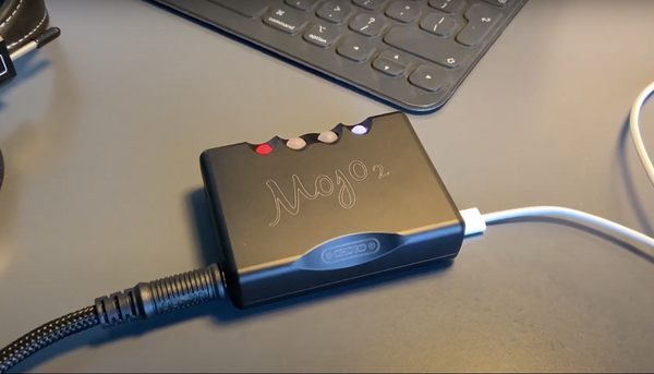 A photo of the Chord Electronics Mojo 2 DAC/Headphone Amplifier, with a thick black cable plugged into the left and a thin white cable plugged into the right.
