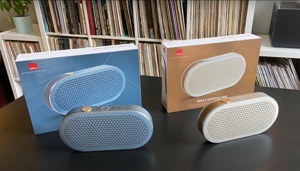A photo of two Dali Katch G2 Portable Bluetooth Speakers, with a blue one on the left and a white one on the right. Behind them are their product boxes.
