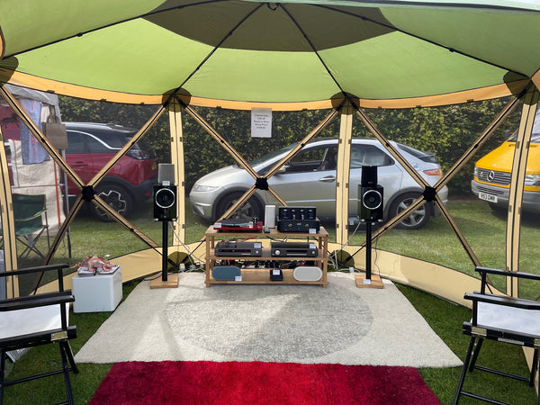 A HiFi system set up in a gazebo tent outdoors. There is a white rug on the ground in the middle of the tent and a red one at the front edge. There are two chairs in the left and right bottom corners.