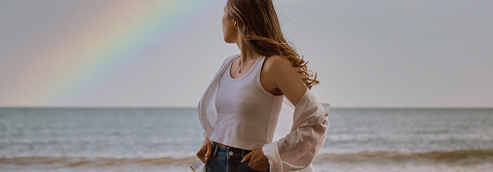 Frau mit langen Haaren steht am Meer und schaut auf einen Regenbogen