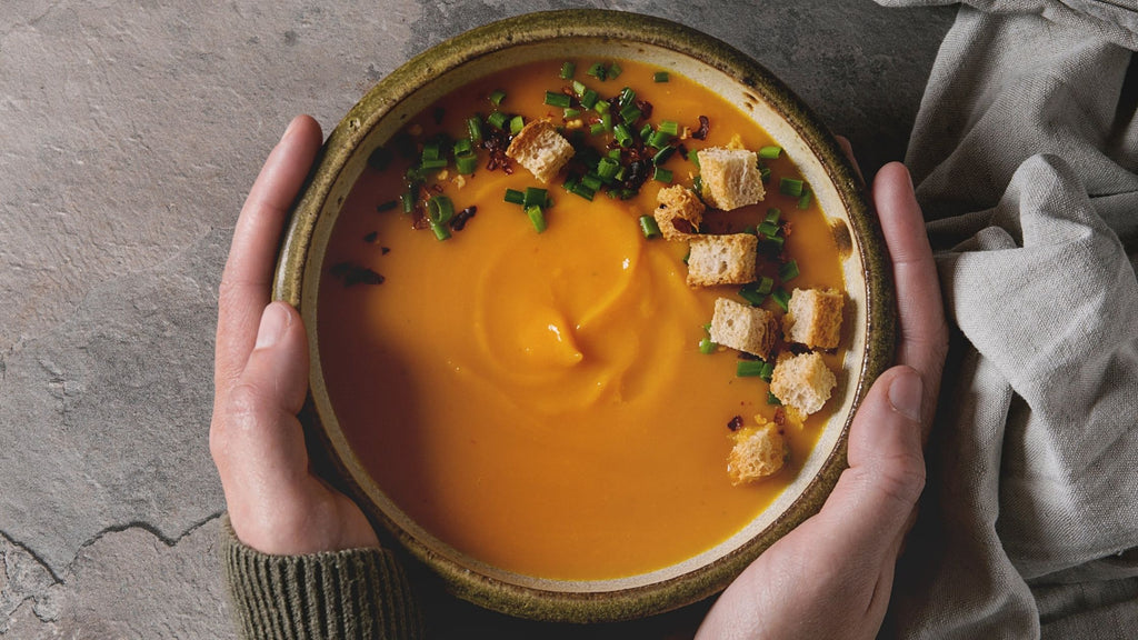 Oppskrift på gulrotsuppe. På bildet ser du en hvit kvinne som holder rundt en skål med gulrotsuppe. Bildet er tatt ovenfra og ned.