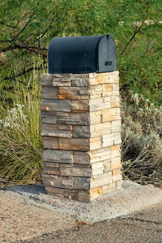 Mailbox post with natural stones in square fashion with black metal container on top in front yard with road nearby and driveway