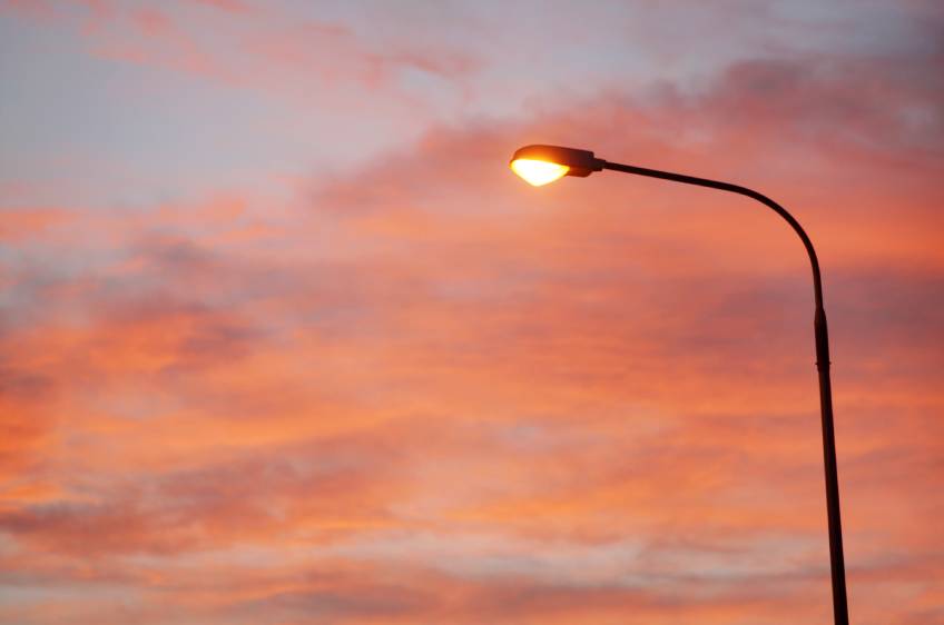 An electric street lamp shines as the sky in the background makes the transition from evening to day.