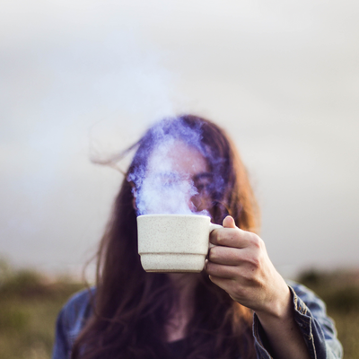 Person with long hair outside holding a steaming cup of nettle tea over face