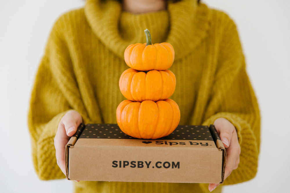 Person holding a Sips by Box with 3 mini pumpkins stacked on top