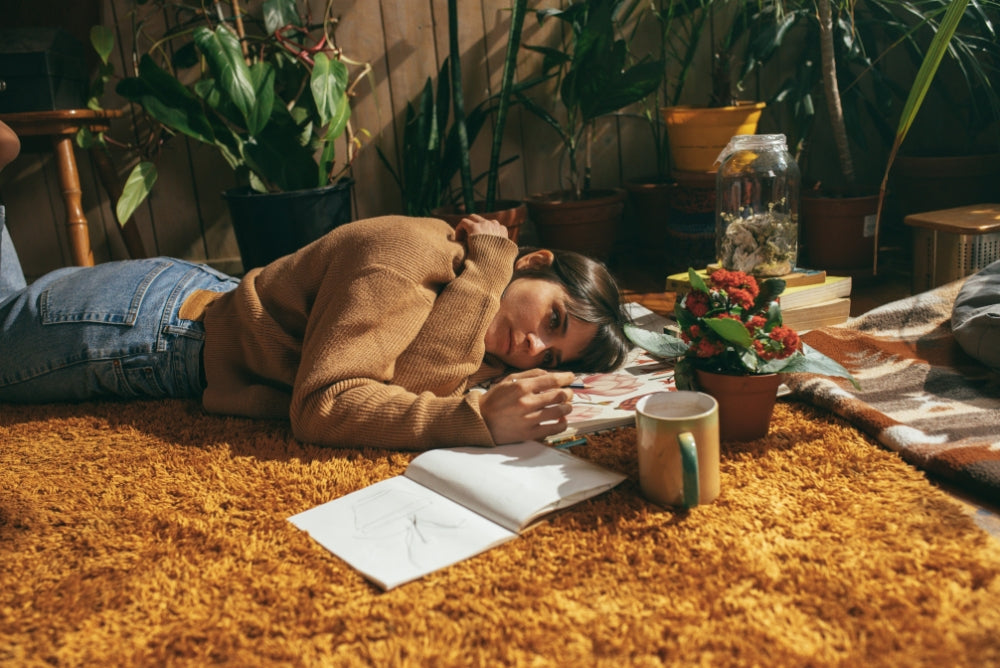 Scene of a woman in an earth toned sweater laying on a shag carpet, writing in a notebook, and drinking a cup of tea