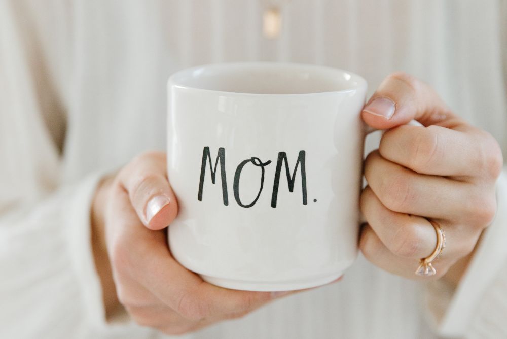 Person in a white shirt with gold jewelry holding a "Mom" Rae Dunn mug