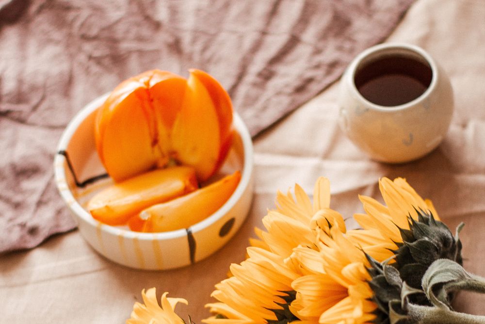 A bowl of peaches, cup of tea, and bouquet of sunflowers on a table with a brown linen tablecloth