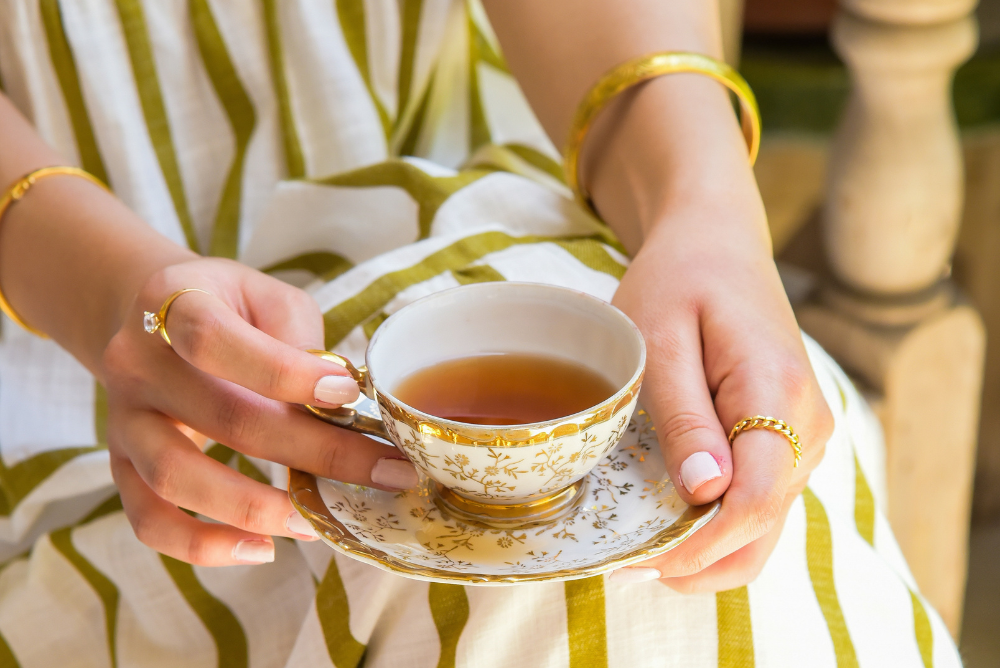 Person in white and green striped dress and gold jewelry holding a gold and white teacup with tea