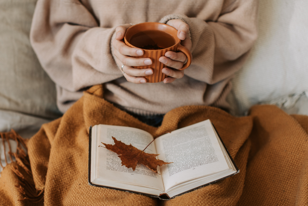 Person in cozy fall sweater with hands wrapped around an orange mug of tea and reading a book with a fall leaf bookmark