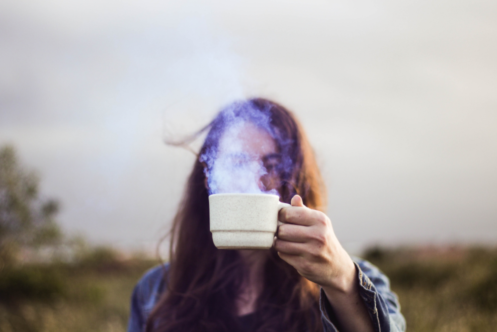 Person with long hair outside holding a steaming cup of nettle tea over face