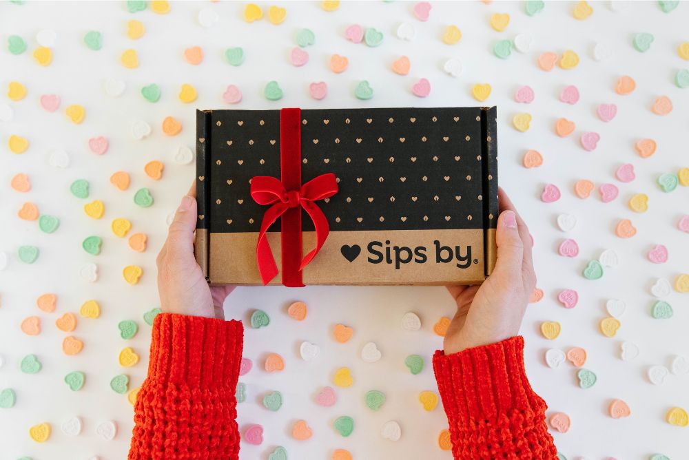 Hands holding up a Sips by Box tea subscription box surrounded by Valentine's Day conversation candy hearts