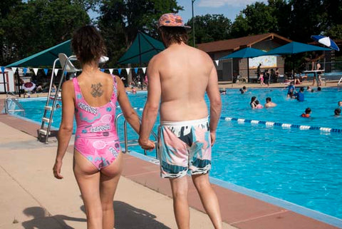 Woman wearing B Fresh All That Racket swimsuit and man wearing B Fresh Cordial swimsuit holding hands by pool.