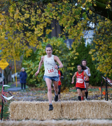 Lauren, a Rose City Track Club member, jumping a hay hurdle during a track event with competitors in the distance.