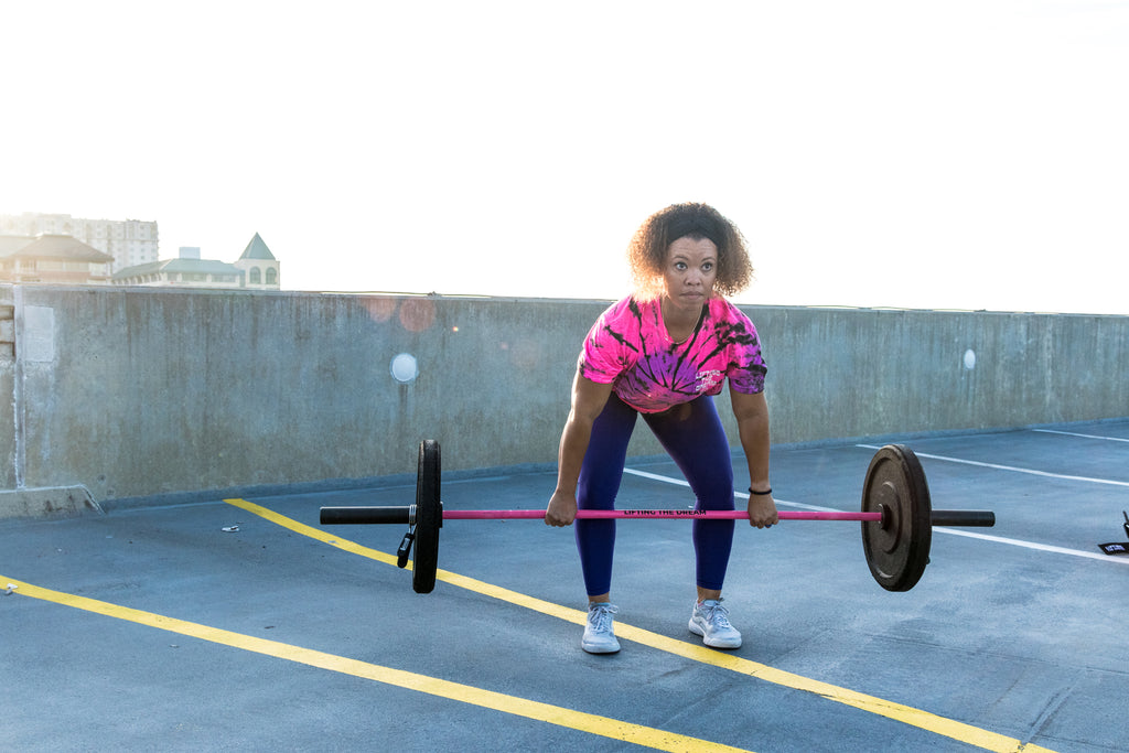 black female athlete lifting a pink barbell in a tie dye shirt on a roof