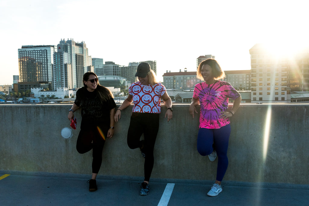Tres atletas femeninas vestidas de entrenamiento sonriendo en lo alto del techo de un estacionamiento al atardecer