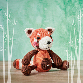 A crocheted red panda with an orange and brown body, white face details and blushing cheeks