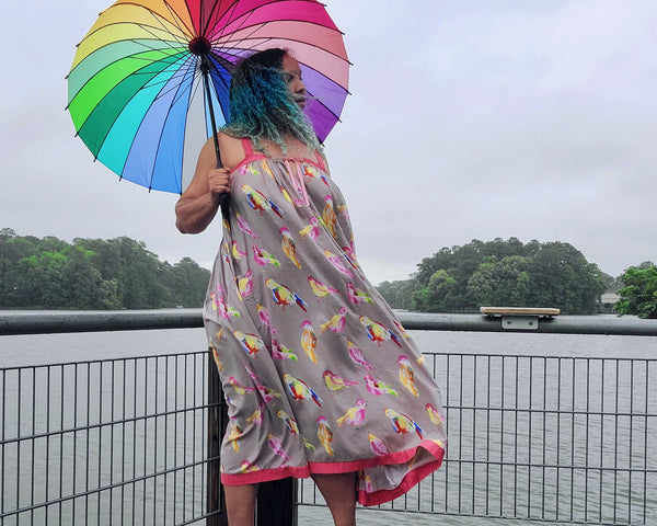 Woman standing outside by a river with a colorful bird print dress and a rainbow umbrella.