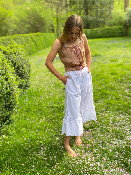 Young woman standing by a boxwood hedge wearing white drawers and a pink camisole.  Her hands are in the plackets