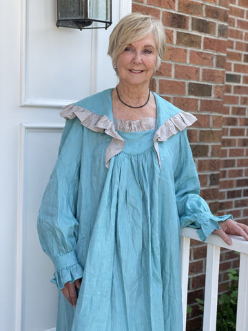 Older woman standing on a brick front stoop wearing a teal linen nightgown.  Her hand is on the white railing.  