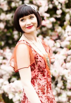 Miss Fisher wearing a red dress with sheer short flutter sleeves, outside and looking over her right shoulder.