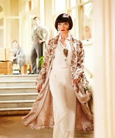 Miss Fisher walking toward camera wearing a silk print swing coat with a white suit undernearth.