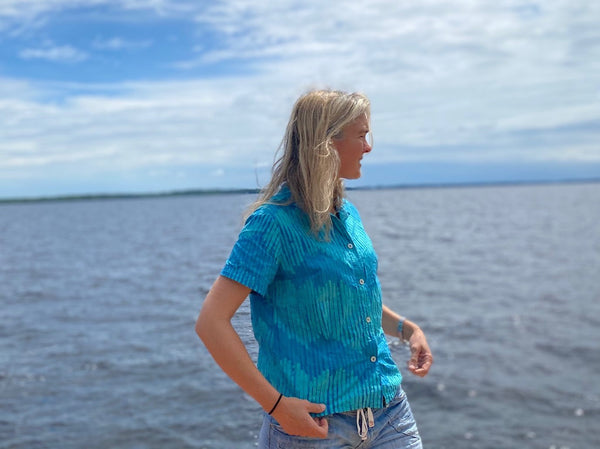 Woman standing by a river wearing a teal block printed button down shirt.