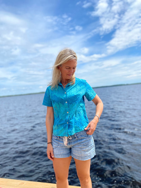 Woman standing by a river wearing a teal block printed button down shirt.