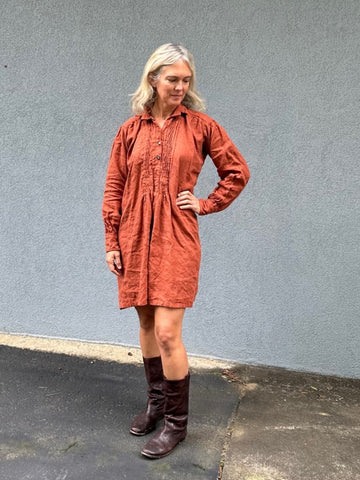 woman standing in a burnt orange linen dress in front of a grey wall with her left hand on her hip.