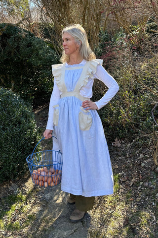 Woman standing outside with hand on her hip, holding a basket of eggs, and wearing a blue and cream apron with ruffled straps.