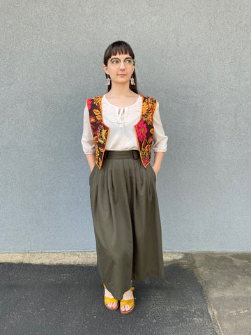 A woman with dark hair is wearing a handmade vest she has sewn. The vest has a dark but colorful floral fabric. She's wearing a green skirt and has her hands in her pockets while she looks to the side.