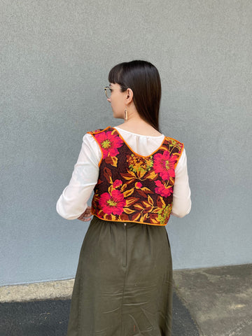 A woman with dark hair is facing away from the viewer, with her back to the camera. She has on a colorful handmade vest and a green skirt. She's wearing a white blouse underneath.