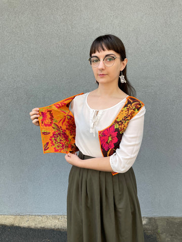 A woman with dark hair is holding open a handmade vest to show off the inside. The vest is reversible, so the outside and inside have different colors to the pattern. She is wearing a white blouse under the vest and a green skirt.