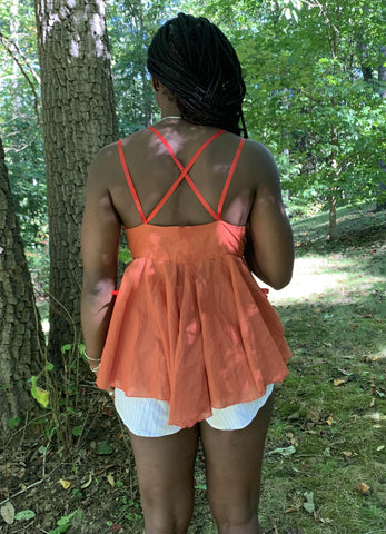 The back of an African American Woman wearing a baby doll top with red crisscross straps. She is standing outside in the woods.
