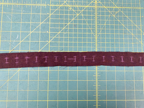 Button holes marked using purple chalk on a strip of dark plum cotton linen fabric.