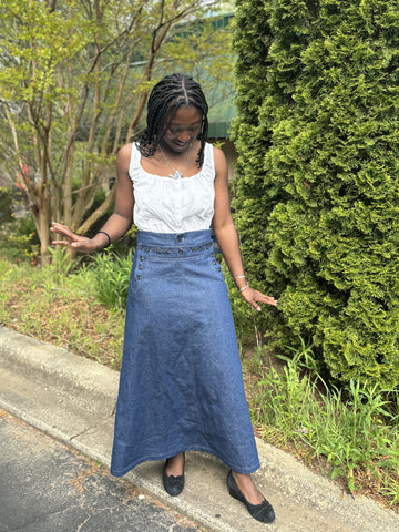 African American woman standing looking down wearing denim sailor skirt with sleeveless white blouse with greenery in the background.