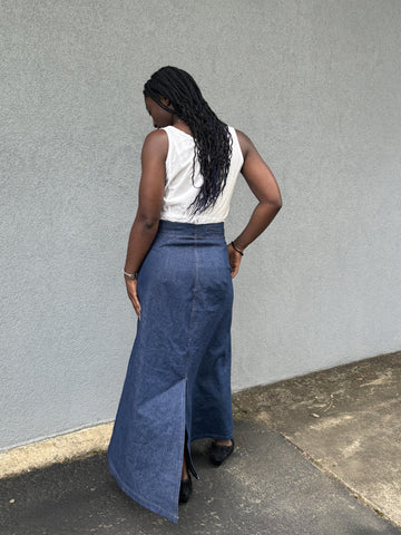 Back of African American woman standing wearing a long denim sailor skirt with a sleeveless white blouse in front of a grey wall outside.