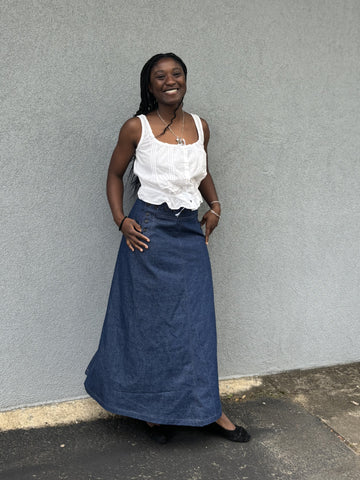 Smiling African American woman standing wearing a long denim sailor skirt with a sleeveless white blouse in front of a grey wall outside.