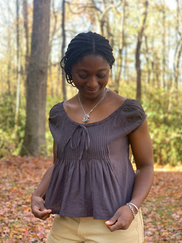African American woman wearing 223 Chemise Blouse looking  down at the garment and smiling. The top is made out of a cotton linen chambray in plum. she is outside with the orange, yellow and red leaves of fall on the ground.