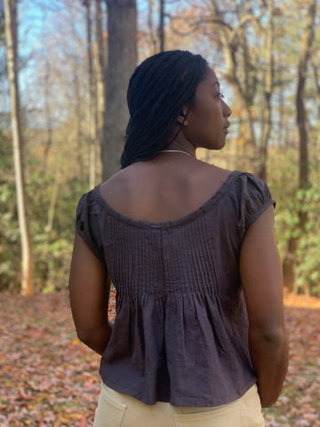 Back of African American woman wearing 223 Chemise Blouse head  looking  towards her right .The top is made out of a cotton linen chambray in plum. she is outside with the orange, yellow and red leaves of fall on the ground.