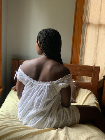 The back of a African American woman sitting on a bed with yellow sheets, wearing 223 A Lady's Chemise in white.