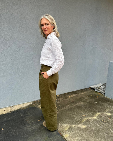 Woman wearing olive colored wide legged trousers and a white linen button down shirt in front of a grey wall outside. Side view.