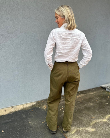 Back view of a woman wearing wide-legged olive colored trousers and a white linen shirt.