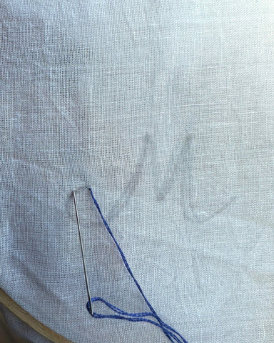 Letter M on white fabric, beginning embroidery with blue thread.